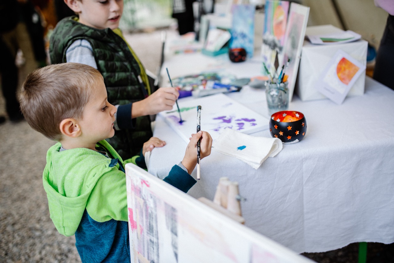 two young boys painting with brushes at a table covered in art supplies and white cloth