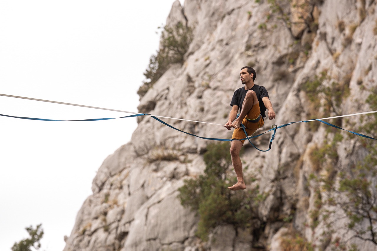 man wearing orange shorts balancing on a slackline high above the ground near rocky cliffs