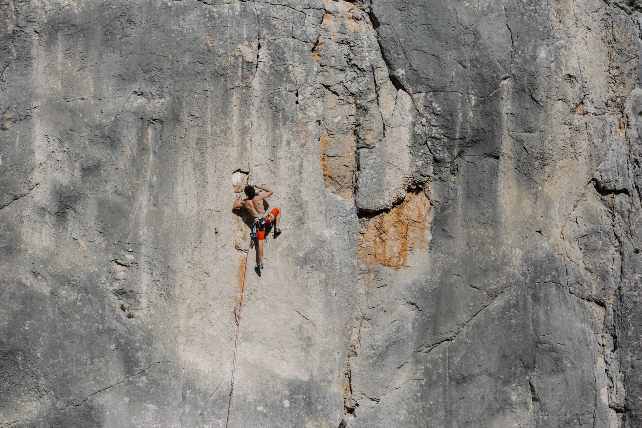 Man climbing a vertical rock face wearing orange shorts and safety harness.