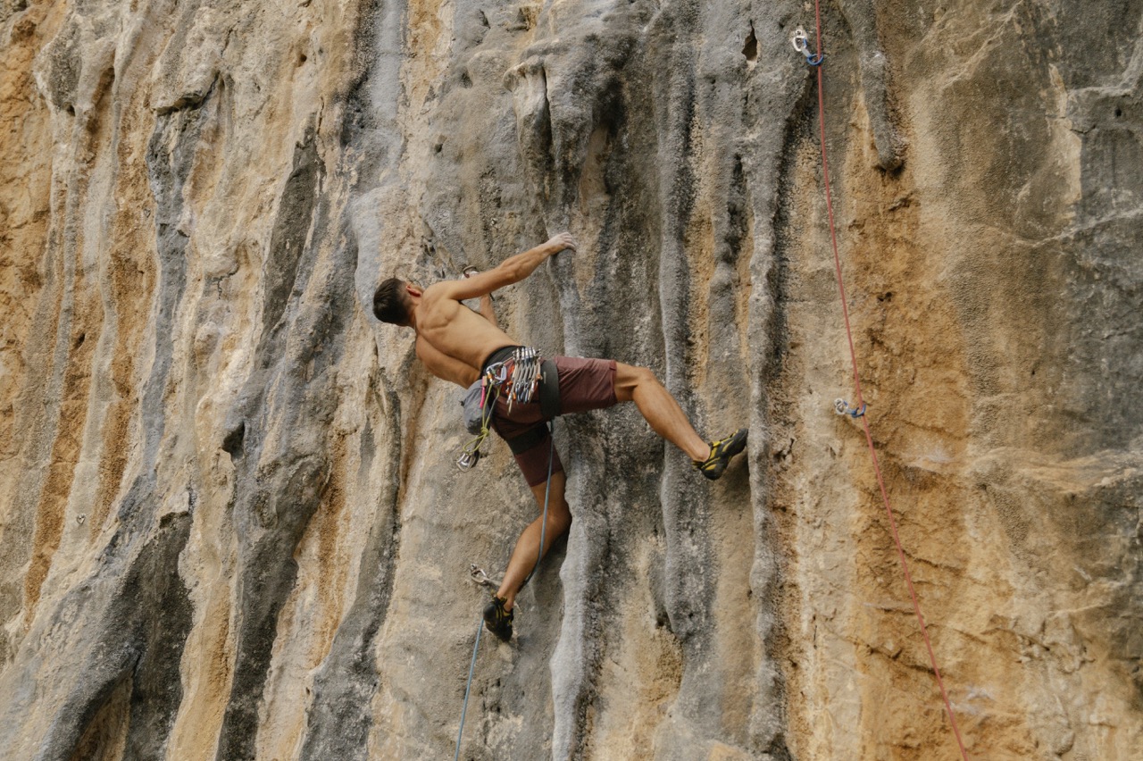 young man rock climbing shirtless on steep textured limestone cliff using safety harness and ropes